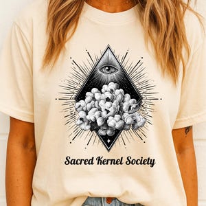 May include: Cream-colored t-shirt featuring a black and white graphic. The design includes an eye within a diamond shape, radiating lines, and a cluster of popcorn. The text "Sacred Kernel Society" is printed below the graphic.