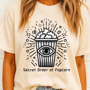 May include: Cream-colored t-shirt featuring a black graphic of a popcorn container with an eye in the center. The popcorn is overflowing, and the design includes radiating lines and the text "Secret Order of Popcorn."