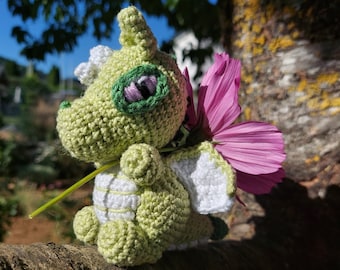 Könnte beinhalten: Ein grüner Häkel-Drache, der eine rosa Blume hält. Der Drache hat weiße Flügel und eine weiße Häkelblume auf dem Kopf.