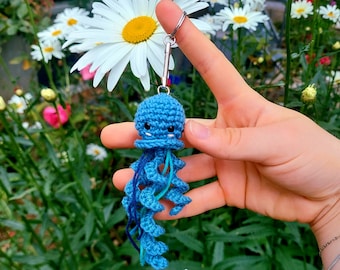 May include: A handmade blue crochet jellyfish keychain with a silver keyring. The jellyfish has a round head with small black eyes and long, curly tentacles in shades of blue. The background features white daisies and green foliage.