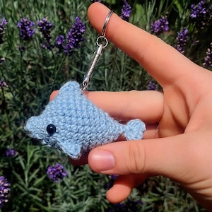 Puede incluir: Un llavero de delfín de crochet azul claro con un ojo negro. El llavero está unido a un anillo de metal plateado.