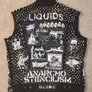 Puede incluir: Chaleco vaquero negro cubierto de tachuelas de metal plateado y parches con logotipos de bandas y las palabras "LIQUIDS", "ANARCHO STENCILISM" y "D.L.I.M.C" en letras blancas. El chaleco tiene una estética punk rock.