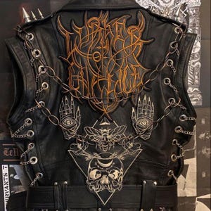 May include: Black leather vest with gold and silver embroidered patches. The vest features a large central design, hand and eye motifs, a skull, and chain detailing. Silver spikes adorn the shoulders, and a belt is present.