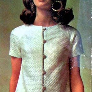 May include: A vintage ivory-colored dress with short sleeves and a textured pattern. The dress features a row of decorative buttons down the front and two small pockets. The dress is knee-length.