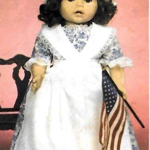 Puede incluir: Una muñeca con un gorro blanco con un lazo azul, un delantal blanco y un vestido con estampado floral azul. La muñeca sostiene una pequeña bandera estadounidense. La muñeca tiene cabello rizado oscuro y está sobre un fondo rosa.