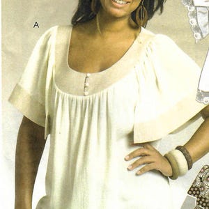 May include: A cream-colored blouse featuring a wide scoop neckline, short flutter sleeves, and a gathered bodice. The garment includes a solid beige trim along the neckline, sleeve edges, and hem, with three small white buttons centered at the chest.