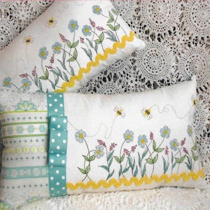 May include: Two rectangular decorative pillows with floral and bee designs. The pillows feature a white background with embroidered blue flowers, green leaves, and yellow zigzag trim. One pillow has a teal polka dot ribbon.