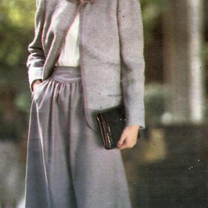 May include: A light grey skirt suit, comprising a jacket with a rounded collar and a knee-length skirt. A black clutch is held. The outfit is worn with a white blouse. The ensemble is a classic business or formal wear.