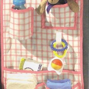 May include: Pink and white checkered car seat organizer with multiple pockets. Includes a baby bottle, teddy bear, diapers, and toys. The organizer is designed to hold baby essentials.