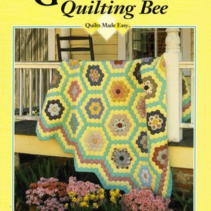 May include: A book cover for "GRANDMA'S Quilting Bee" with the subtitle "Quilts Made Easy." The image shows a colourful quilt draped over a porch railing. The quilt has a yellow, blue, and pink colour scheme. Floral arrangements are in the foreground. The book's title is in large black letters.