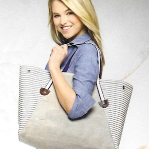 May include: A large tote bag with a beige canvas body and a striped top section. The bag has white straps with a dark stripe and brown leather accents. The bag is being held by a person.