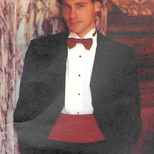 Peut inclure: Un smoking noir avec une chemise blanche, un nœud papillon bordeaux et une ceinture de smoking bordeaux. La veste de smoking a un revers cranté. La chemise a des boutons noirs. Le fond est un mur en marbre.
