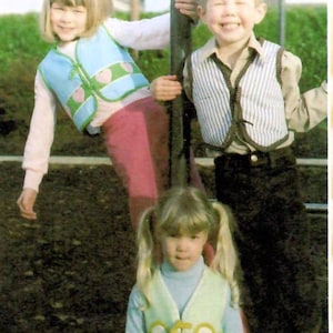 May include: Three children in vests pose for a photo. The children are wearing blue and green vests, decorated with heart shapes and other designs. The children are smiling and appear to be enjoying themselves.