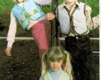 May include: Three children wearing vests pose for a photo. The children are wearing vests in shades of blue and green, with heart-shaped and other decorative elements. The children are smiling and appear to be enjoying themselves.