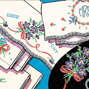 May include: A collection of white fabric linens featuring colourful embroidered floral motifs, lace trim edges, and text including MINE, GOOD NIGHT, and MRS. The designs incorporate red ribbons, blue flowers, and birds, presented against a bright blue background.