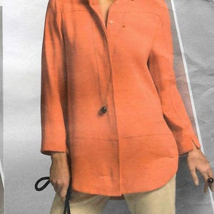 May include: A bright orange long-sleeved button-down shirt with a collar and a single visible dark button at the waist, paired with light beige trousers. The shirt features a horizontal seam across the chest and a curved hemline at the bottom.