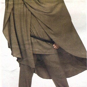 May include: A taupe-colored ensemble consisting of a high-neck tunic, straight-leg trousers, and a wide, draped poncho-style wrap. The outfit is accented with a thin black belt at the waist and a wide, metallic gold-toned cuff bracelet on one wrist.