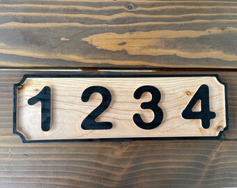 Pode incluir: Uma placa de número de casa de madeira com números pretos 1234. A placa tem uma borda preta e uma forma ligeiramente arredondada.