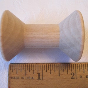 Puede incluir: Una bobina de madera con un centro cil&iacute;ndrico y extremos c&oacute;nicos. La bobina es de madera de color claro y mide aproximadamente 5 cm de largo. Una regla de madera est&aacute; en primer plano.
