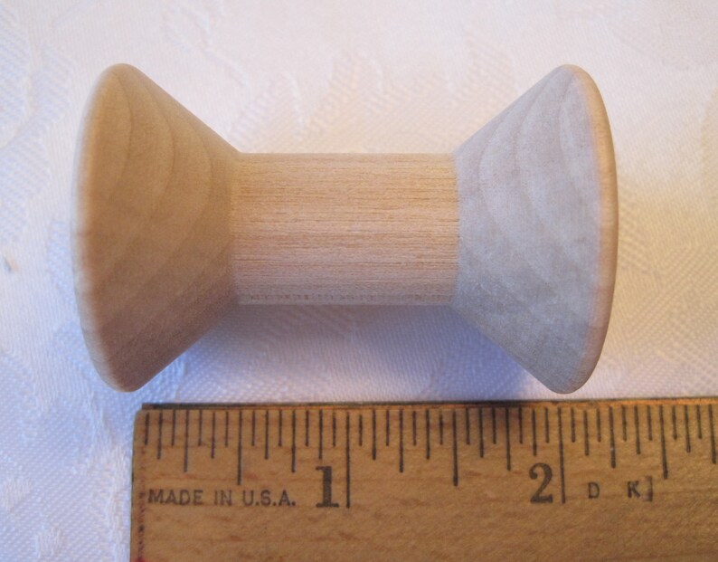 Puede incluir: Una bobina de madera con un centro cil&iacute;ndrico y extremos c&oacute;nicos. La bobina es de madera de color claro y mide aproximadamente 5 cm de largo. Una regla de madera est&aacute; en primer plano.