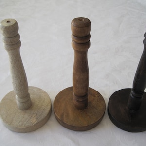May include: Three wooden paper towel holders in varying shades of brown. The holders have a round base and a central post with a knob at the top. The colors range from light beige to dark brown.