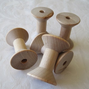 May include: A collection of six unfinished wooden spools. Each spool has a wide, flat base and top with a cylindrical center. The wood has a light, natural color, and the spools are arranged on a white, textured surface.