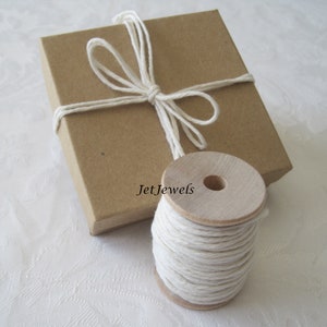 May include: A small, brown cardboard box tied with white twine, next to a wooden spool of white string. The box is closed and the twine is tied in a bow. The spool of string is full and ready to use.