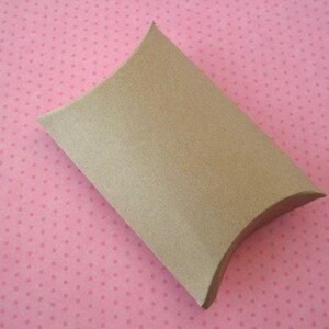 May include: A brown cardboard pillow box with a curved shape. The box is on a pink background with white polka dots.