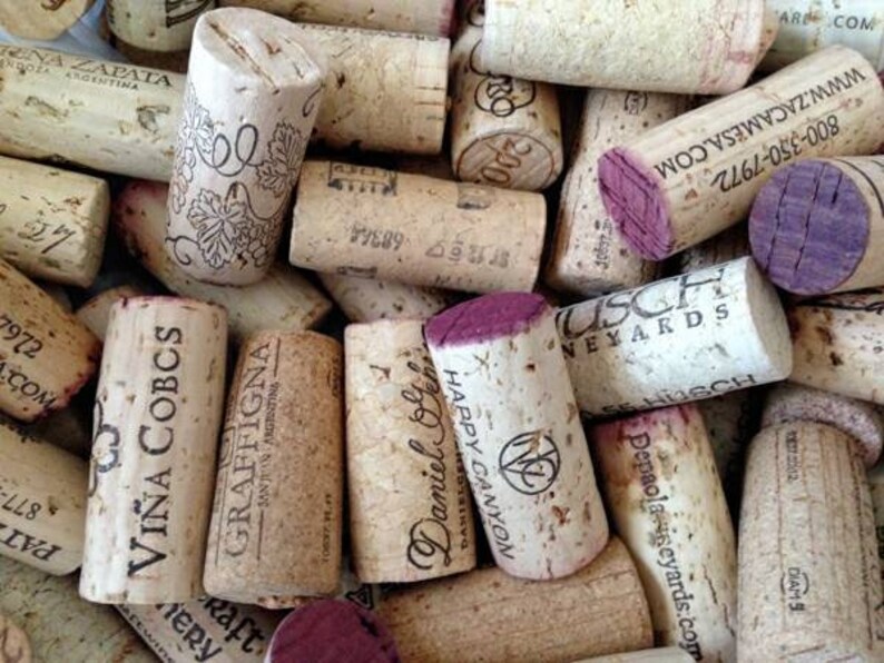 May include: A collection of used wine corks, featuring various brands and designs. The corks are primarily beige with some having purple-stained ends. Text and logos are printed on the corks, showcasing wineries and vineyards.