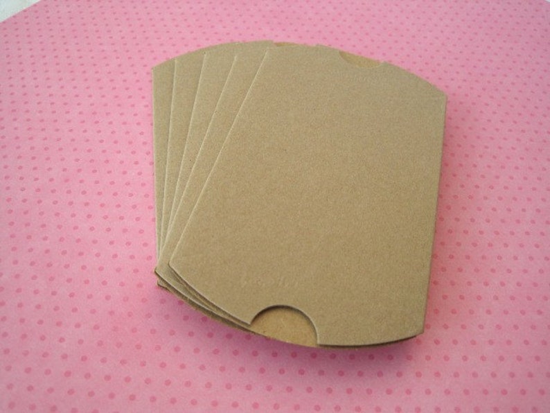 May include: A stack of five beige, die-cut paperboard drink sleeves. Each sleeve has a curved top and a small cutout on one side. The sleeves are arranged on a pink background with small, evenly spaced, darker pink polka dots.