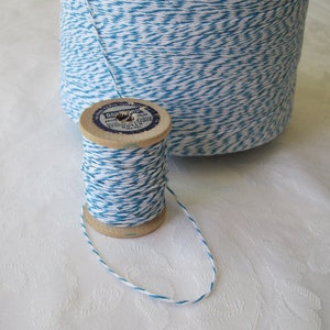 May include: Two spools of blue and white baker's twine. One spool is a large cylinder, and the other is a smaller wooden spool. The twine is a twisted pattern of white and turquoise. The wooden spool has a blue and white label.