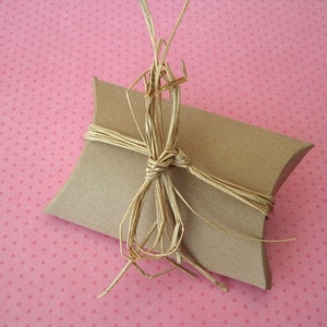 May include: A small, brown, pillow-shaped gift box tied with natural raffia ribbon. The box is made of cardboard and sits on a pink background with small white polka dots. The raffia is tied in a bow.