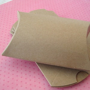 May include: A stack of brown, pillow-shaped gift boxes. The boxes are made of a light brown cardboard material and have curved edges. The boxes are stacked on a pink surface with small white polka dots.