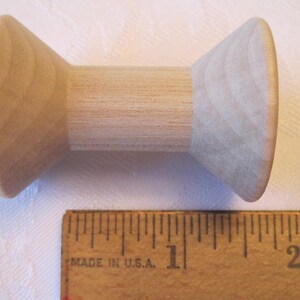 Puede incluir: Una bobina de madera clara con un centro cil&iacute;ndrico y extremos c&oacute;nicos. Una regla de madera con marcas en pulgadas se coloca junto a la bobina. La bobina mide unos 6 cm de largo.
