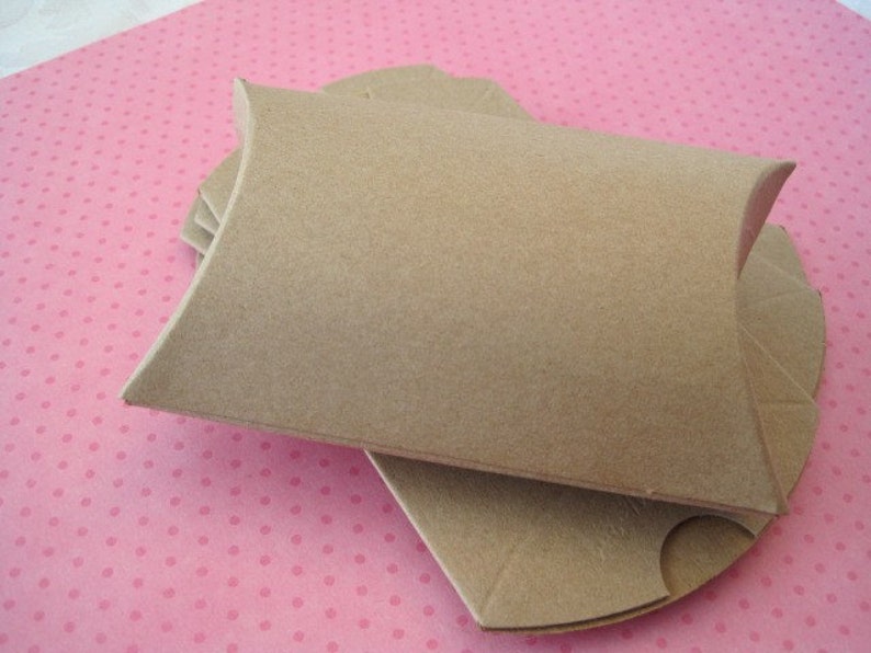 May include: A stack of brown pillow boxes, made of cardboard, are arranged on a pink polka dot surface. The boxes have a curved shape with folded edges, designed to hold small items. The boxes are empty and ready for use.