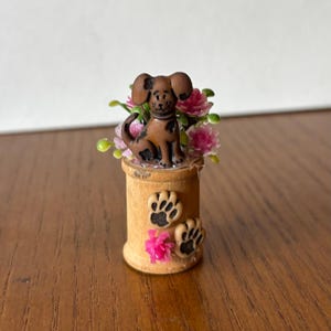 May include: A brown dog figurine with black spots sits atop a wooden spool, surrounded by pink flowers and green leaves. Two paw prints and pink flowers are on the spool. The figurine has large ears and a smiling face.