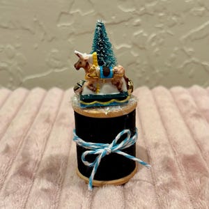 May include: A miniature Christmas carousel figurine with a brown horse, a blue and gold saddle, and a small green Christmas tree. The figurine sits atop a black spool of thread tied with blue and white twine.