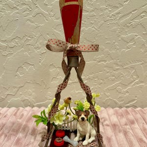 Peut inclure: Un tampon décoratif avec un manche rouge et un ruban à motifs marron et blanc. Le tampon présente une figurine de petit chien, un os et deux bols rouges. Le chien est assis devant une clôture blanche avec des fleurs.
