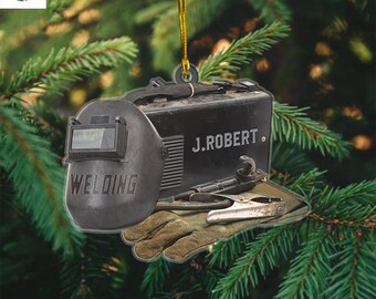 May include: A black and gray ornament featuring a welding helmet, toolbox, gloves, and tools. The word "WELDING" is printed on the helmet. The toolbox has the name "J. ROBERT" printed on it. A gold string is attached for hanging.