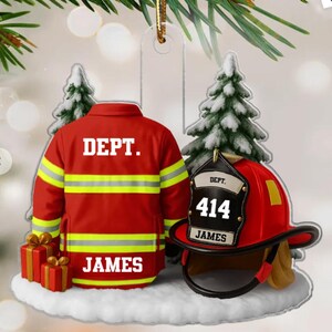 May include: A Christmas ornament featuring a firefighter's uniform and helmet. The red jacket has yellow stripes and the word "DEPT." and "JAMES" in white. The red helmet has the number "414" and the name "JAMES". Two small gift boxes and a snowy base complete the design.