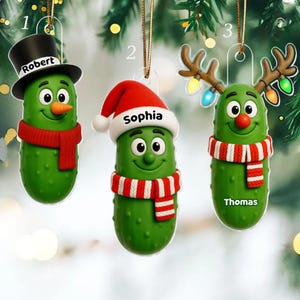 以下が含まれることがあります： クリスマスツリーに飾られた3つのピクルスのオーナメント。各緑色のピクルスは笑顔で、帽子やスカーフを身に着けています。オーナメントには、Robert、Sophia、Thomasの名前がパーソナライズされています。