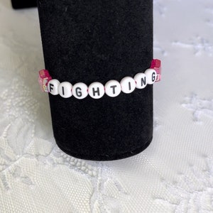 May include: A pink and white beaded bracelet with the word "FIGHTING" spelled out in black letters.