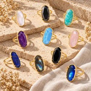 May include: A collection of ten gold-toned rings, each featuring a large, faceted, oval-shaped gemstone in various colors including white, black, turquoise, purple, light blue, pink, deep blue, iridescent gray, and dark blue.