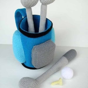 Puede incluir: Una bolsa de golf de crochet con dos palos de golf, una pelota de golf y un tee. La bolsa es azul y gris, y los palos son grises.