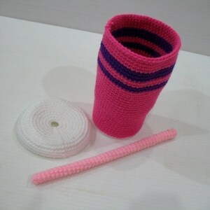 May include: A pink crocheted cup with purple stripes, a white crocheted lid, and a pink crocheted straw.