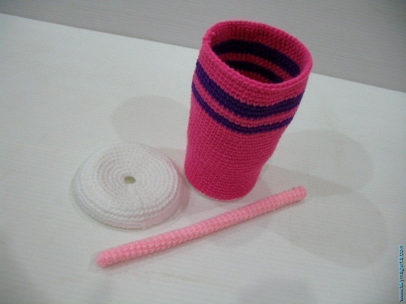 May include: A pink crocheted cup with purple stripes, a white crocheted lid, and a pink crocheted straw.