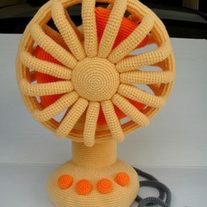 Puede incluir: Un ventilador de crochet con una base amarilla y aspas naranjas. El ventilador tiene un cable con un enchufe negro y una pequeña araña de crochet.