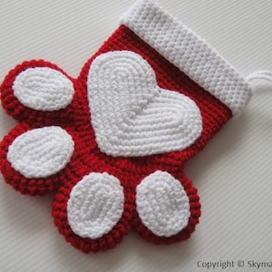 May include: Red and white crocheted Christmas stocking shaped like a paw print with a white heart in the center.
