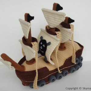 May include: A crocheted pirate ship toy made with brown, beige and grey yarn. The ship has three sails, a skull and crossbones and a wooden mast.