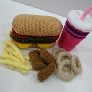 May include: A crocheted hamburger, french fries, onion rings, and a pink and white crocheted soda cup with a pink straw. The food items are all made of yarn and are a fun and whimsical way to represent a classic fast food meal.